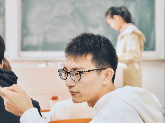 Dans une salle de classe, un jeune garçon, assis à son bureau, se tourne vers son camarade de classe et semble lui parler de manière agressive